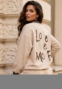 Eine Frau mit langen, welligen Haaren präsentiert die beigefarbene Bomberjacke „Love More“ im Freien und hebt dabei den auffälligen Schriftzug auf dem Rücken hervor. Diese stilvolle Bomberjacke bietet eine vielseitige und bequeme Passform und eignet sich perfekt zum Kombinieren zu jeder Jahreszeit. Das einzigartige Design und die motivierende Botschaft machen sie zur idealen Wahl für modebewusste Kunden, die sowohl nach Trends als auch nach Inspiration suchen. Ein Gebäude mit strukturierter Steinfassade im Hintergrund verleiht dem Look einen urbanen Charme und unterstreicht die Alltagstauglichkeit des Outfits.