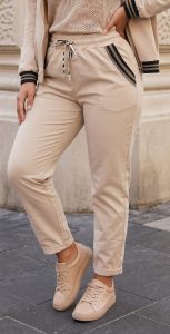 Komfort-Hose „Easy Chic“ – beigefarbene Jogginghose mit stilvollen, schwarz gestreiften Taschen, die am Unterkörper des Models zu sehen sind, kombiniert mit einer passenden Jacke und beigen Sneakern. Aus weichem, bequemem Material gefertigt, ideal für den Alltag; perfekt für vielseitige Styling-Möglichkeiten und ganztägigen Tragekomfort. Abgebildet in einer Außenkulisse vor einer eleganten Steinmauer, um den raffinierten und zugleich lässigen Look hervorzuheben.