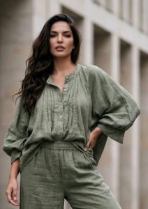 Eine Frau mit langen dunklen Haaren steht im Freien und trägt die olivgrüne Baumwollbluse „Natural Ease“ sowie eine dazu passende Hose, wodurch sie einen entspannten, modernen Look präsentiert. Die atmungsaktive Baumwollbluse bietet den ganzen Tag über Tragekomfort und einen lässigen Stil – perfekt für den vielseitigen Alltag. Sie posiert mit einer Hand in der Hüfte und blickt selbstbewusst in die Kamera. Dabei unterstreicht sie die elegante Passform und das zeitlose Design, die dieses Set zu einem Must-have für Kundinnen machen, die Komfort und Raffinesse suchen.