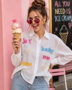 Eine Frau in einer weißen „Lucky Five“-Hemdbluse mit buntem Aufdruck und einer stylischen Sonnenbrille; ein fröhlicher Sommerlook – perfekt für einen Ausflug zum Eisessen.