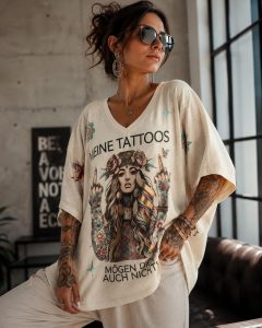 Eine tätowierte Frau präsentiert das locker sitzende „Love my Tattoos“-Shirt, das sich durch einen einzigartigen deutschen Text und eine auffällige Illustration einer tätowierten Frau mit Blumenverzierung auszeichnet – perfekt, um Individualität zum Ausdruck zu bringen und Körperkunst zu feiern. Dieses T-Shirt wurde mit Blick auf Komfort und Stil entworfen und eignet sich ideal als Freizeitkleidung oder als Statement-Stück. Im Hintergrund sind große Fenster zu sehen, durch die natürliches Licht hereinfällt, sowie ein Wandschild, das die authentische Innenkulisse unterstreicht.
